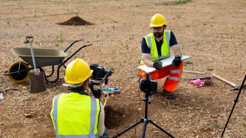 AncientCraft/Emma Jones Two people are wearing yellow hi-vis jackets and hard hats. Both are crouching on the ground and are surrounded by different equipment, including tools and a wheelbarrow. 