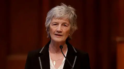 PA Media Shows a women with grey hair, a black jacket and white top, standing at a mic