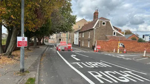 A street blocked off by signs saying no entry, with an arrow advising drivers to turn right. there are houses along the right hand side of the road