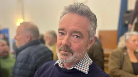A man with grey hair and a grey beard, wearing a dark jumper, looks at the camera