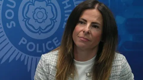 Proffitt is positioned slightly off-center and is facing toward the right side of the frame. She has long, straight hair that falls past the shoulders, with a darker shade at the top transitioning into a lighter tone toward the ends. She is wearing a light-colored, textured jacket with a subtle check or woven pattern. Underneath the jacket, she is wearing a plain, high-neck top. The jacket appears to have decorative details near the collar area, including small round elements resembling buttons or embellishments.