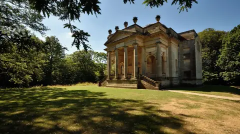 A grand, stone Greek-style building with four pillars at the front. There are large archways at the front and two staircases on either side of the entrance. On top of the roof are five stone sculptures which look like large urns. There is a patch of grass in front of the building and trees surrounding it. The sun is casting shadows of the trees on the grass and the sky is bright blue.