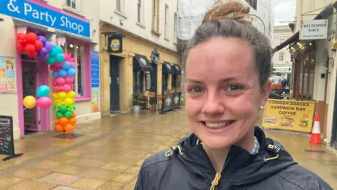 A woman with brown hair in a top knot smiles on a wet street lined with shops and restaurants on a rainy day. She is wearing a black raincoat with a yellow zip that has been zipped to her neck and is wearing pearl stud earrings. A colourful balloon arch can be seen at the entrance of a party shop.