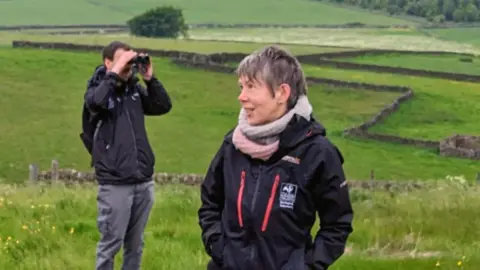 Sheffield and Rotherham Wildlife Trust A woman with short grey hair is wearing a black coat and pink and grey scarf, She is standing in a field looking to one side and there is a man in the background looking through a pair of binoculars