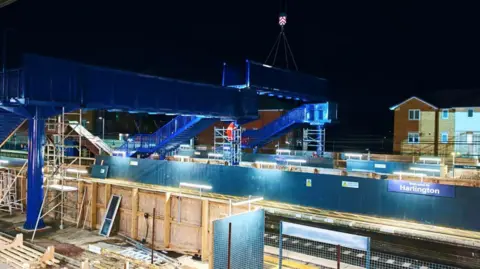 Network Rail A crane lifting a section of a blue footbridge into place at Harlington train station at night time. There are wooden hoardings on the platforms.