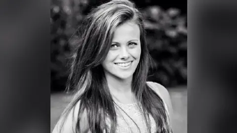 Chipchase family A black and white image of India Chipchase, with long dark hair, looking at the camera and smiling. She is wearing a short-sleeved top. A tree is behind her. 