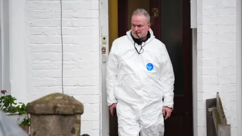 PA Media A police officer leaving the home, a white-painted terrace with a brown wood front door and geraniums in a window box. He is wearing a white all-in-one boiler suit and is chewing on the arm of his glasses while looking thoughtfully grim.