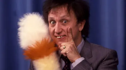 BBC Ken stands with his trademark feather duster in hand in front of a draped curtain. His hand rests on his face. He's wearing a suit.