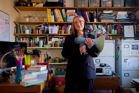 Laurence Cawley/BBC Professor Scott holding a megaphone. She is wearing a blue suit and glasses and is surrounded by shelves of books, two filing cabinets and some brain wave monitoring equipment on a table