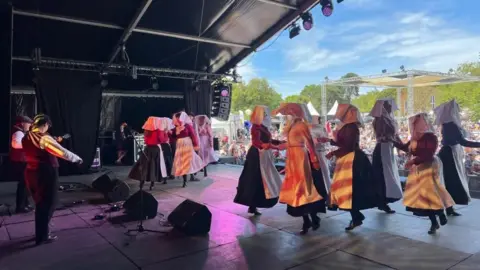 Screen Cornwall A stage full of dancers who are skipping in a circle with old-fashioned clothing on. Two people can be seen playing the guitar at the back of the stage, and the image is taken from the back of the stage, looking out towards the dancers and the crowd.