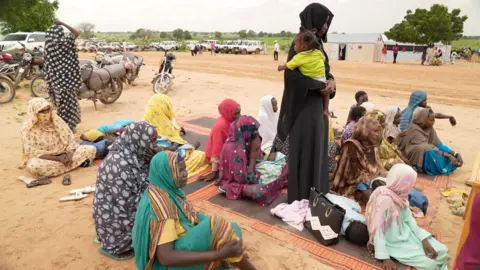 Kevin McGregor / BBC Para wanita duduk di atas tikar di tanah di sebuah kamp di Adré 