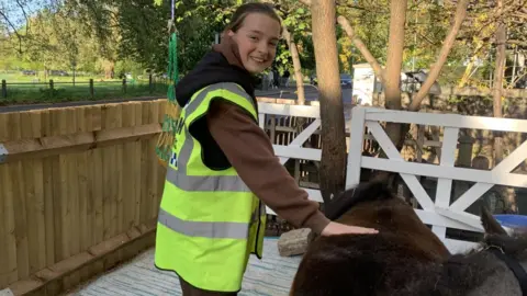 A teenage girl with a hi-vis jacket on petting a short brown pony