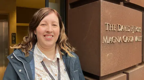 Alan Webber, BBC A woman with blonde hair, wearing a cream and orange blouse and a blue jacket, smiles at the camera. She is standing in front of a building with a large marble-style sign, which has gold letters reading; The David PJ Ross Magna Carta Vault.