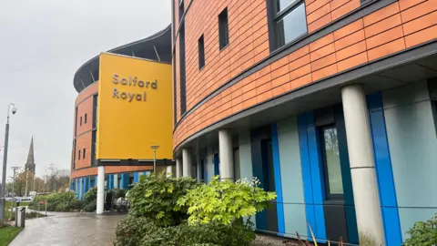 Photograph of Salford Royal. The image of the hospital includes a yellow sign, tiles on the building, a pathway and shrubs.