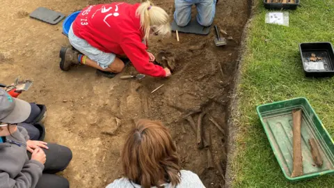 A woman wearing a red top is on her hand and knees scraping dirt from a skull while three other people kneel in the trench. There are other bones visible.