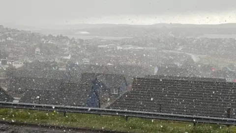 Snow falling across Lerwick in Shetland. Looking out from the top of a hill over lots of houses with black roofs. The sea and hills can be seen in the distance. A dark sky. 