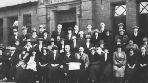 DT Architectural Design A black and white image showing a large group of people stood or sat together in rows. Many are wearing hats and are seemingly dressed in formal clothing. 