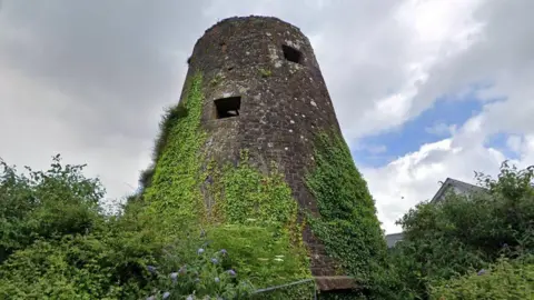 A large circular stone structure seen with square windows and a flat roof. It has vegetation growing up it and trees either side. 