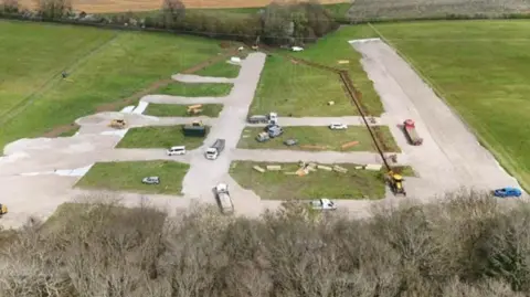 Flamstead Parish Council An aerial shot of a green field edged by a row of trees at the bottom edge. Concrete hard standing has been laid in a grid pattern and a trench has been dug through middle of the site. There are vans, diggers and cars moving around the site. 