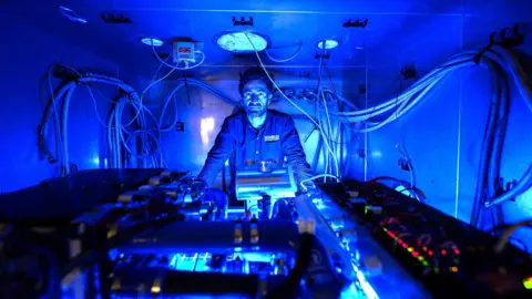 OceansLab A heavily blue image of a man in overalls in a small metallic room with a hydrogen engine in front of him.