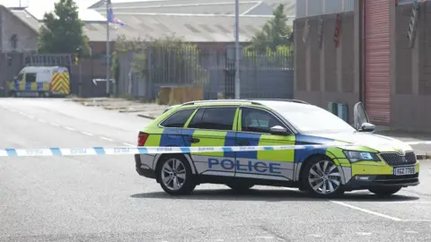 A yellow, blue and white police car is behind police tape that is cordoning off a road. A police van is parked in the background. The road is empty. 