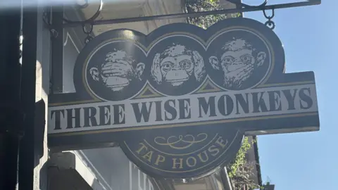 A close up of the Three Wise Monkey pub's sign outside the pub. Three monkey heads are on the sign each with a different expression. 