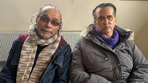 A man and woman sitting in front of a cream wall. There is a radiator behind them and they are both looking into the camera. The man is wearing rectangular tinted glasses, a cream scarf and denim jacket. The woman is wearing a purple turtleneck jumper and grey padded coat fur around the hood. 