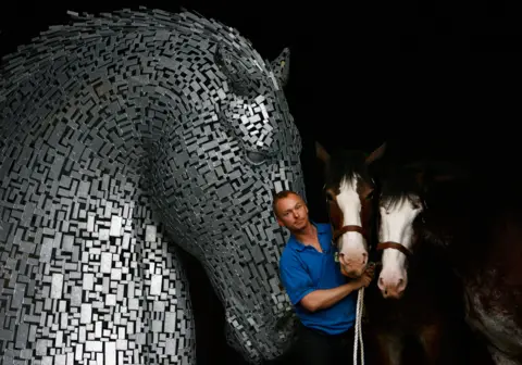 Getty Images Sculptor Andy Scott wearing a blue t-shirt and holding the reins of two Clydesdale horses, stands next to one of his sculptures of the Kelpies