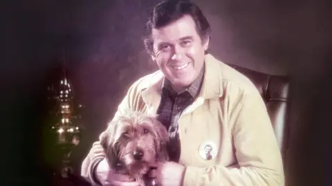 A man in a yellow jacket beside a parafin lamp with a dog on his lap