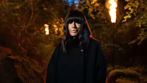 BBC/Studio Lambert Claudia Winkleman dressed in a black cloak outside with fire on large lamps burning behind her. 