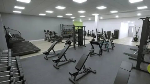 A computer generated image showing what the new gym looks like. It is a large room with dark grey padded flooring covering the weight lifting area, and vinyl flooring near the treadmills and exercise bikes. The walls are a light grey with white overhead lighting.