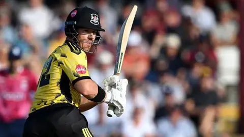 D'Arcy Short, wearing a black helmet, yellow shirt, black trousers, black pads and white gloves, plays a cricket shot while holding his bat