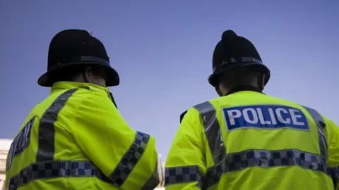 Stock image taken from the back of two police officers stood side by side wearing fluorescent police jackets and black police hats.