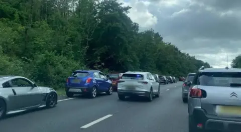 An unmarked police car with its blue lights on drives along the A38 in Devon to the scene of a crash. Both lanes of traffic have moved over to allow space for the vehicle to move through.