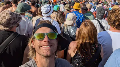 Lewis Simonds Lewis Simonds, smiling at the camera, with a cap on his head, to the side, sunglasses on, in front of a large crowd of people, at Glastonbury