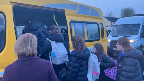 A number of school children are seen wearing black and navy coats and school bags are seen boarding a yellow school bus.