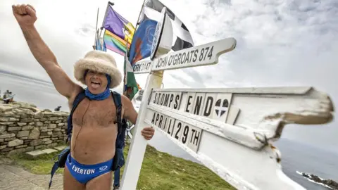 Story Picture Agency/Shutterstock Speedo Mick wearing his blue Speedos, a woolly hat, scarf, backback and nothing else, with one arm around a white wooden direction sign at Land's End, and the other arm in the air and his mouth smiling and open. Pictured in 2023.