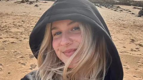 Danni Diston takes a selfie as she is standing on a sandy beach. She has long blonde hair, blue eyes and a small silver nose ring. She is wearing a black hoodie with the hood up. Behind her are a couple of seaweed-covered rocks.