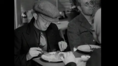 British Pathe A black and white news clip of a man eating a meal at a table with a knife and fork. He is wearing a black overcoat, a dark shirt and a lighter coloured tie. He is also wearing a tweed-style checked cap and looking down at his food. A man to his left has an empty plate in front of him and is looking off into the distance.