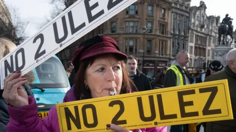 PA Media A woman is holding a sign saying no to ULEZ and has a whistle in her mouth 