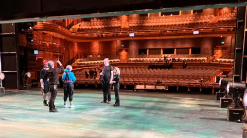 Five members of the WNO production crew stand on a large stage, lit in blues and creams. Two of the crew are wearing hard hats and one is pulling a cable towards a cast member's back, who has a strap on her back. In the background is the Wales Millennium Centre's auditorium with the lights up. Members of the production crew are stood watching. 