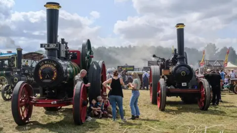 "Two large vintage steam traction engines with tall black chimneys and red wheels are displayed at an outdoor heritage rally. Visitors stand and sit between the engines on a grassy field, while steam drifts across the background under a partly cloudy sky."
