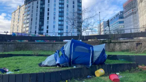 A tent pitched on a verge of grass in the middle of the Bearpit roundabout in Bristol. There are buckets and rubbish scattered on the ground. Tall buildings can be seen in the background.