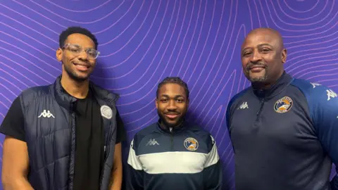 (Left to right) Earl Burgess, Wilfred Santhe and Dean Blake. Three men in dark blue sports clothing are smiling at the camera and stood against a purple swirly background
