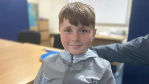 Carson is wearing a grey jacket. He has light brown hair and is smiling at the camera.