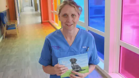 Nurse Lin Fidgin stands in a hospital ward with brightly coloured windows and holds up a children's book which has a title called Bess of Clara Vale. She has blonde hair which is tied back and is wearing blue nurse scrubs.