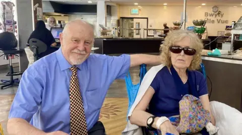 Spinal Research An older couple sat side by side in a cafe. The man is bald with a white beard and mousache and is wearing a blue shirt and brown check tie. He is smiling and looking at the camera, with one hand round the back of his wife, who is sat in a wheelchair. She has blond hair and is wearing a blue t shirt and sunglasses with a colourful bag on her lap.