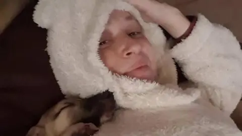 Nia McGregor Nia lying down wearing a hooded white fluffy jumper with her hand up to her hood. Her dog is resting on her shoulder sleeping.