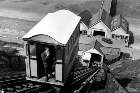 Getty Images A black and white image of Jack Brownless, maintenance manager of C. Horne & Co LTD testing the Saltburn Cliff Railway. He is standing in the open door of the carriage as it rides along the track on the cliffside. There are three small buildings at the base and a pier behind that. There is another carriage on the parallel track.