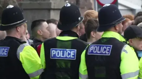 A picture of football fans crowded together in a group. They are stood behind a line of police officers. They are wearing green hi-vis vests on top of their clothes.